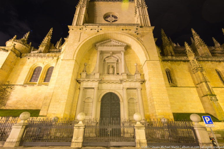Upward night view of Segovia Cathedral’s monumental arched entrance, with a smaller doorway, iron fence, and tower masonry above.