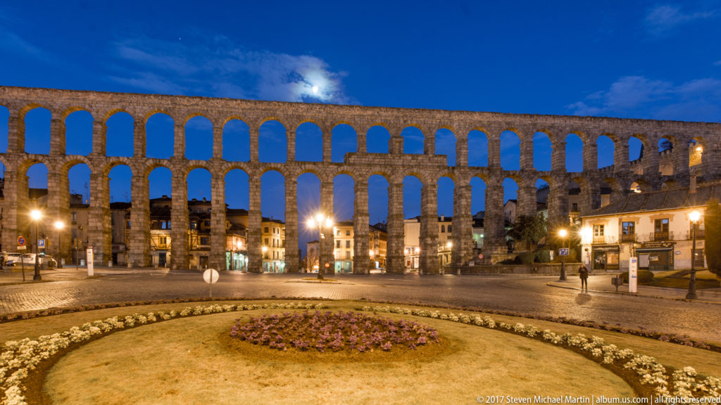 Segovia’s Roman aqueduct crosses a public square at blue hour, with illuminated granite arches above a circular landscaped bed.