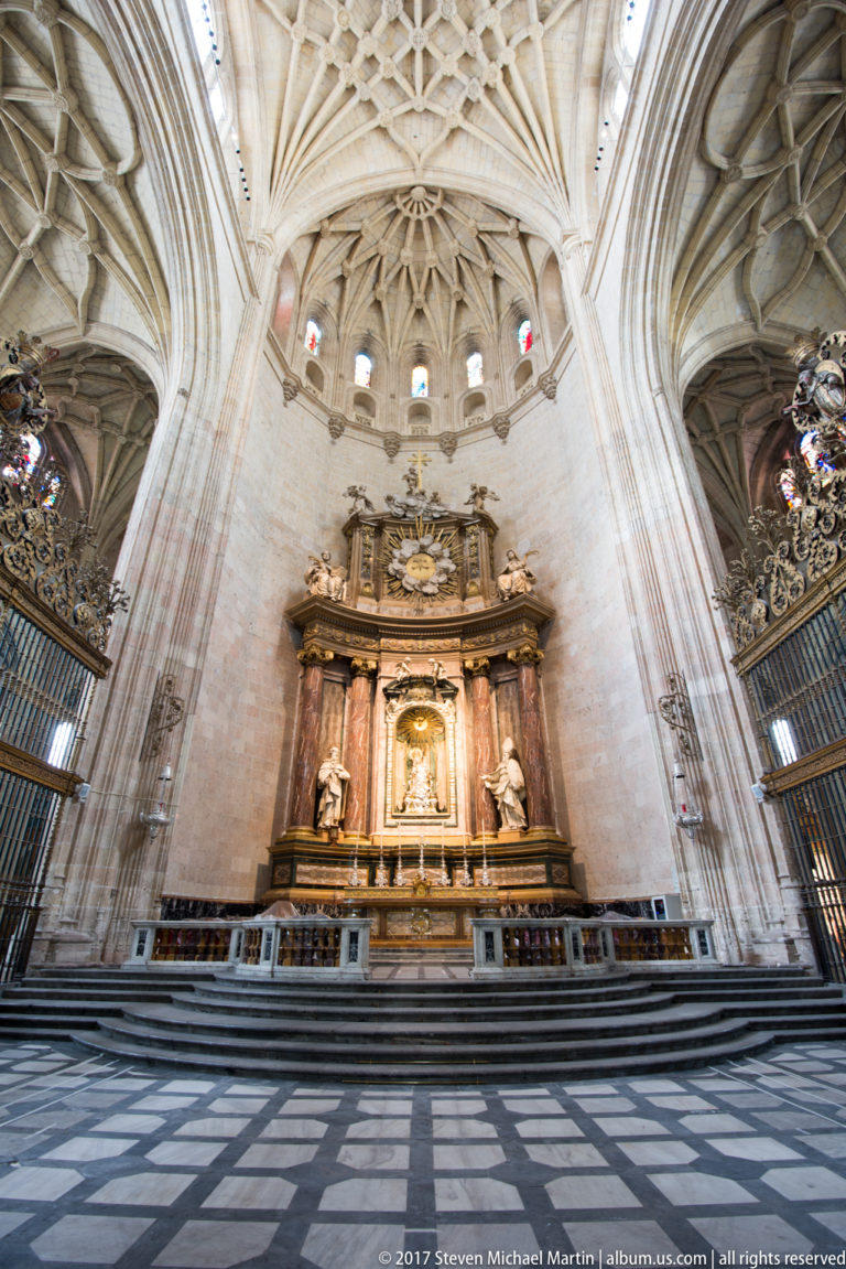Gilded chapel altarpiece beneath elaborate ribbed stone vaulting, with patterned floor and metal screens inside Segovia Cathedral.