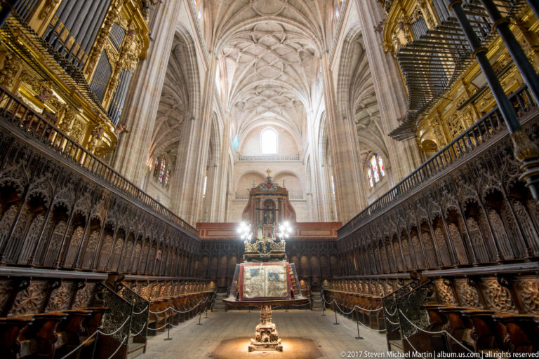 Symmetrical cathedral choir with carved wooden stalls on both sides, central lectern, gilded details, organ, and high ribbed vaults.