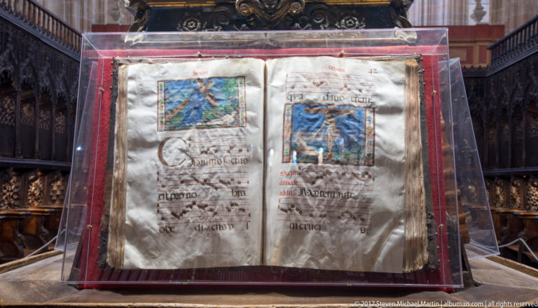 Open illuminated manuscript in a clear display case, with handwritten text, red rubrication, and two colorful miniatures.