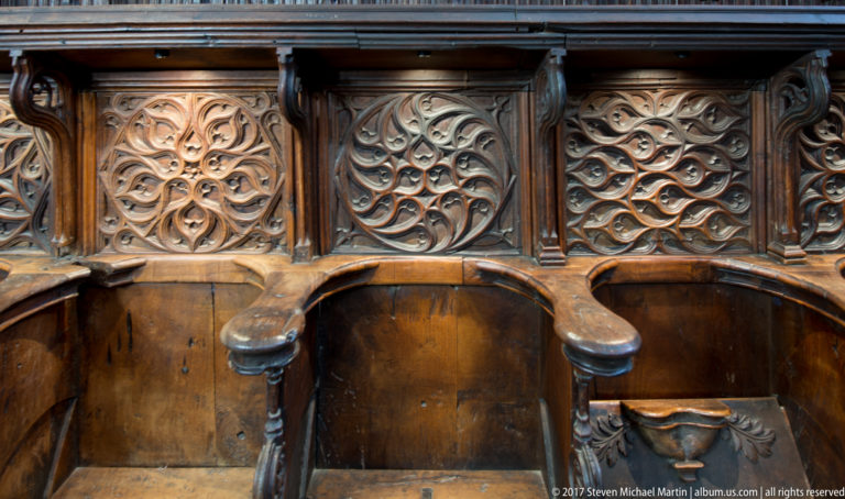 Close-up of dark wooden choir seats with curved armrests and intricate carved openwork panels of interlacing motifs.