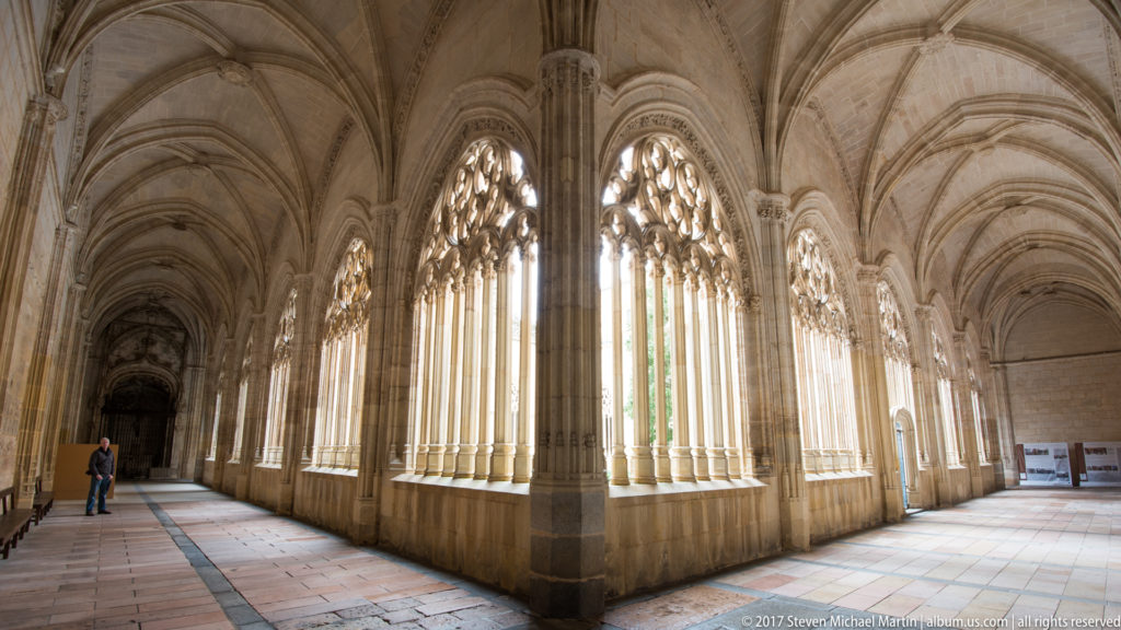 Gothic cloister gallery with tall traceried openings, ribbed vaults, stone piers, and sunlit tiled walkway at Segovia Cathedral.