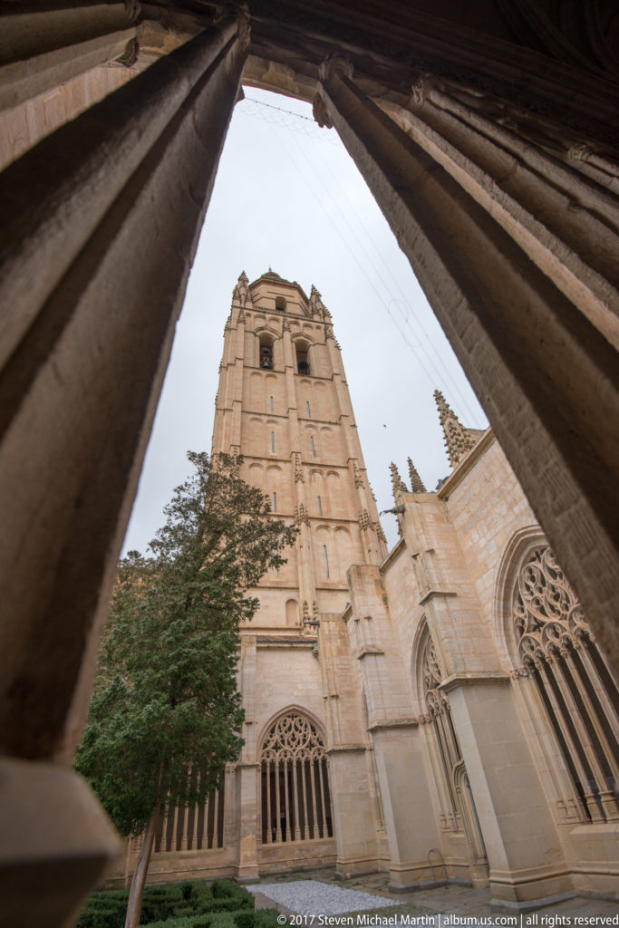 Segovia Cathedral tower and Gothic windows seen through the dark stone tracery of a cloister opening.