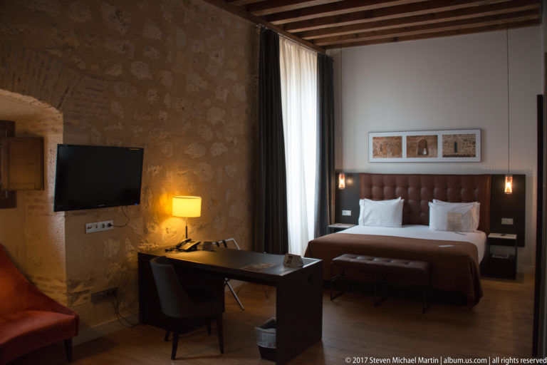 Guest room with exposed stone wall, timber ceiling, bed, desk, television, bench, lamps, and tall curtained window.