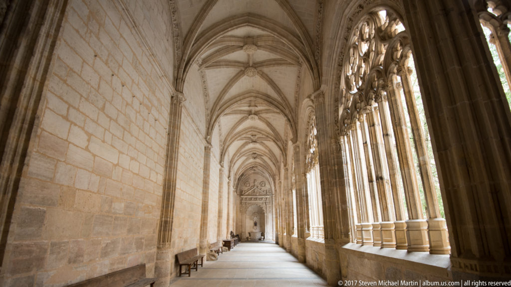 Long Segovia Cathedral cloister corridor with ribbed vaults, stone benches, and tall traceried openings along the courtyard side.