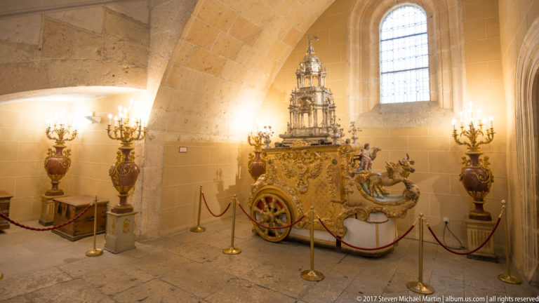 Ornate gilded ceremonial carriage with large wheels, sculptural decoration, rope barriers, urns, and branching lights inside Segovia Cathedral.