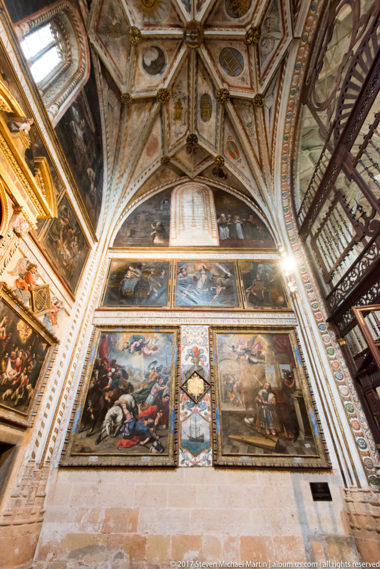 Upward view of two large religious canvases, smaller painted scenes, and a richly decorated ribbed chapel vault.