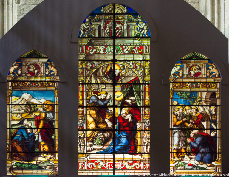 Three arched stained-glass windows with colorful religious figures and narrative scenes, the central lancet taller than the two flanking windows.