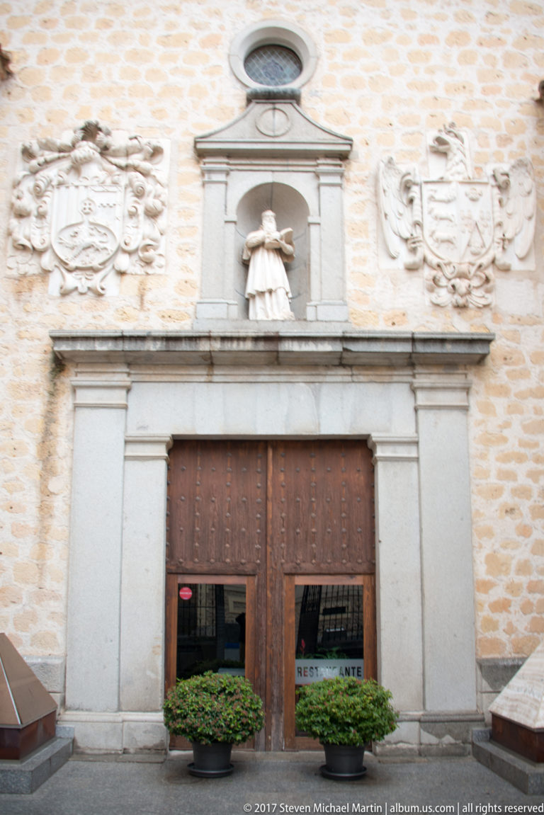 Stone portal with studded wooden doors, central sculptural niche, circular opening, and large carved heraldic shields on both sides.