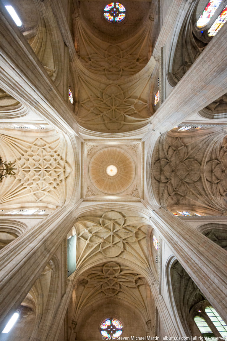 Upward view of pale stone cathedral piers meeting elaborate ribbed vaults, with small stained-glass windows around the upper walls.