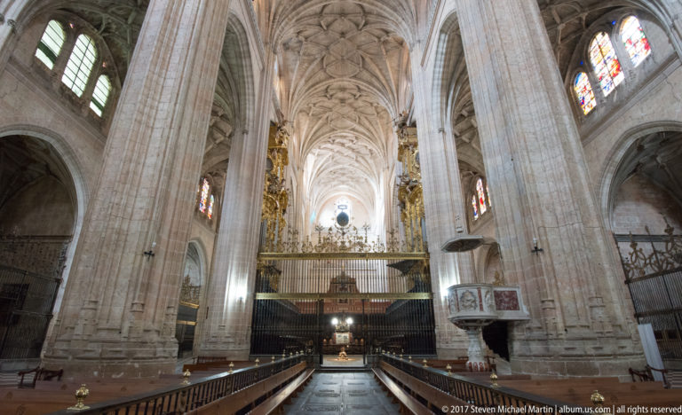 Central aisle of Segovia Cathedral with tall stone piers, ribbed vaults, wooden seating, screened choir area, and pulpit.
