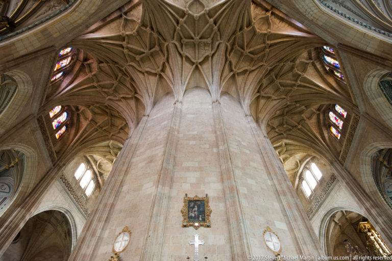 Tall pale stone pier branching into complex ribbed vaults, with stained-glass windows and a small framed devotional image below.