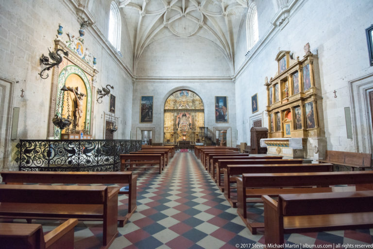 Bright cathedral side chapel with wooden benches, patterned floor, wall altarpieces, paintings, and a wrought-metal enclosure at left.