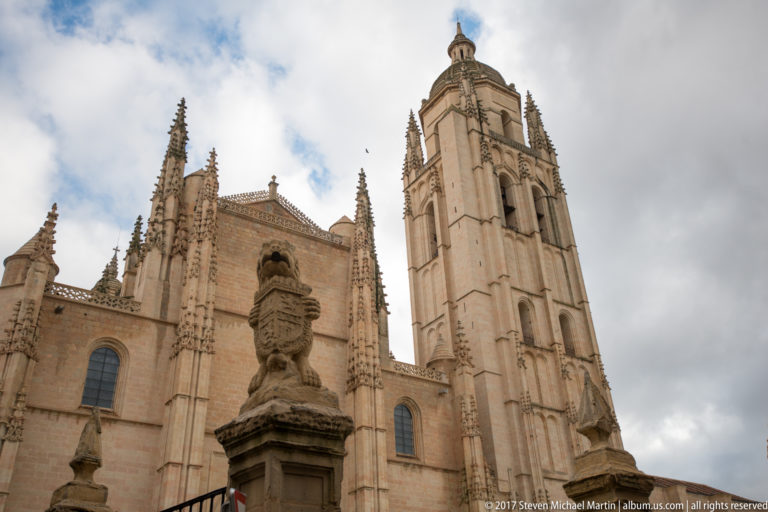 Low-angle exterior view of Segovia Cathedral with its tall bell tower, pinnacles, arched windows, and a stone finial in front.