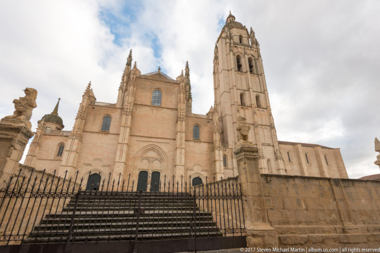 Segovia Cathedral beyond an iron fence, showing broad stone elevations, stepped approach, pinnacles, arched windows, and tall bell tower.