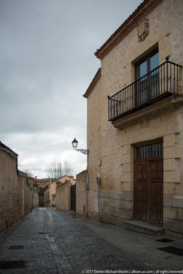 Cobbled lane beside a two-story stone building with iron balcony, wooden door, crest, and projecting street lamp.