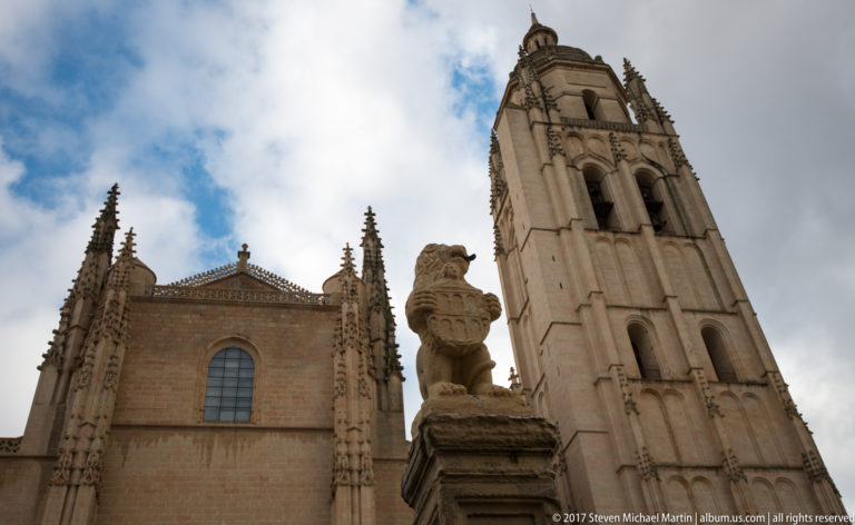Low-angle view of Segovia Cathedral bell tower above a carved stone finial, with belfry openings and pinnacles against clouds.