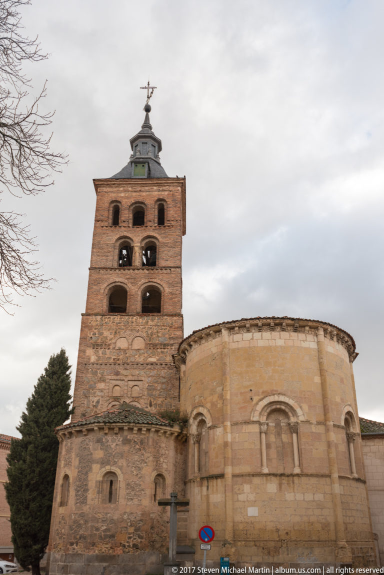 Romanesque stone apse beside a tall square brick bell tower with stacked arched openings and decorative blind arcades.
