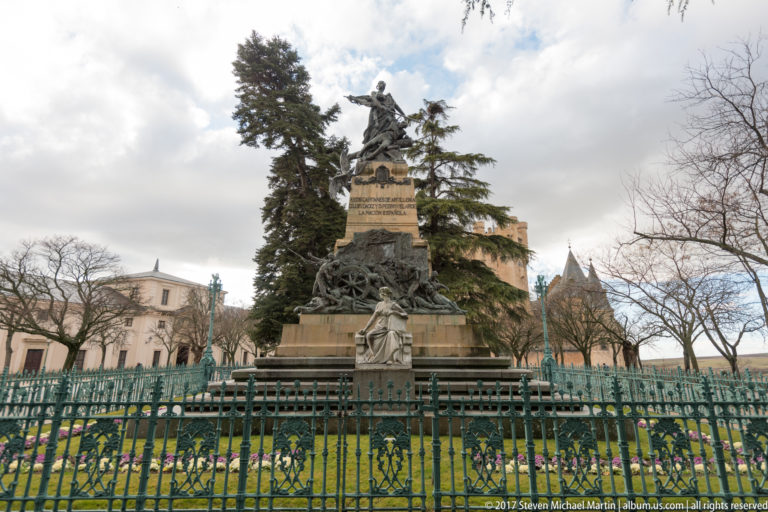 Bronze multi-figure monument on a tall stone pedestal inside a fenced garden with trees and flower beds.