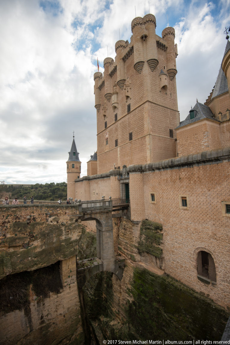 Oblique exterior view of the Alcázar of Segovia with turreted keep, masonry walls, slate roofs, and rocky defensive edge.