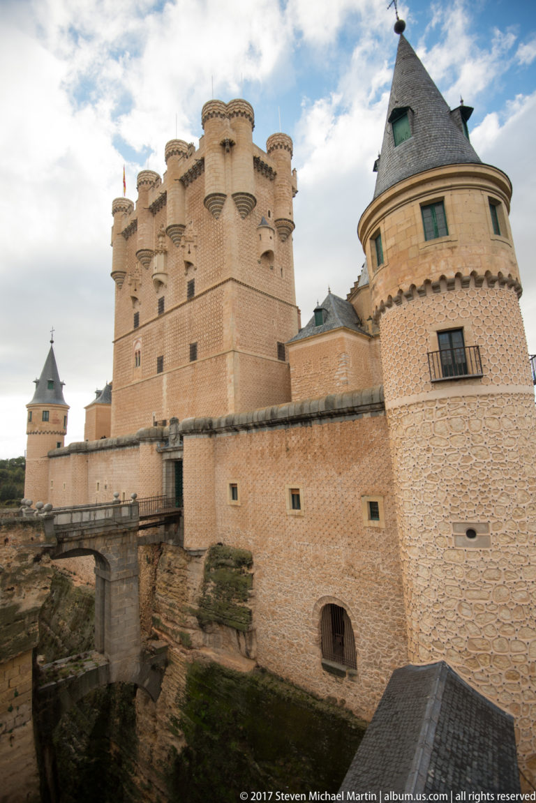 Alcázar of Segovia keep behind a rounded conical-roofed tower and warm masonry outer wall above a rocky edge.