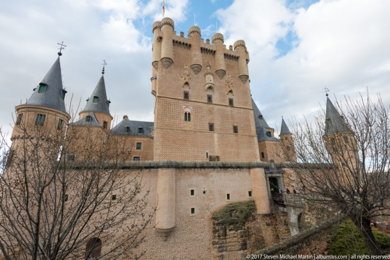Tall crenellated Alcázar tower with corner turrets, smaller slate-roofed towers, bare branches, and rocky ground below the walls.