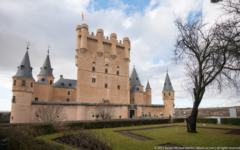 Alcázar of Segovia beyond a formal lawn, with central keep, conical slate-roofed turrets, warm walls, and a leafless tree.