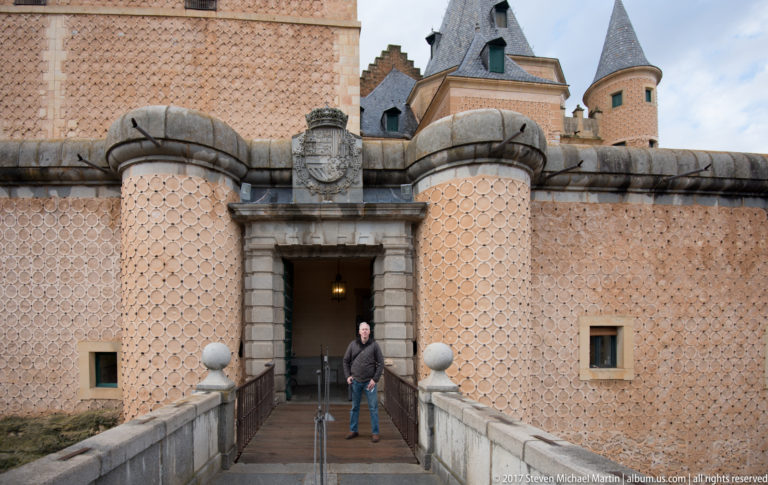 Stone bridge leading to the Alcázar entrance, with patterned walls, rounded projections, heraldic shield, towers, and one visitor.