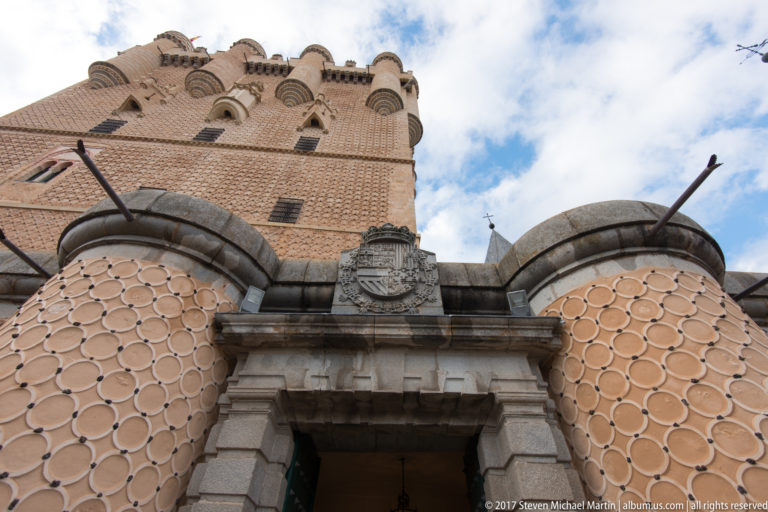 Close low-angle view of the Alcázar entrance with carved heraldic shield, stone portal, circular-patterned wall decoration, and keep above.