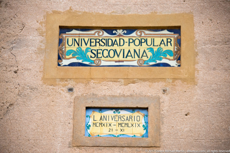Two decorative ceramic plaques on a wall, including a large sign reading Universidad Popular Segoviana and a smaller anniversary plaque.