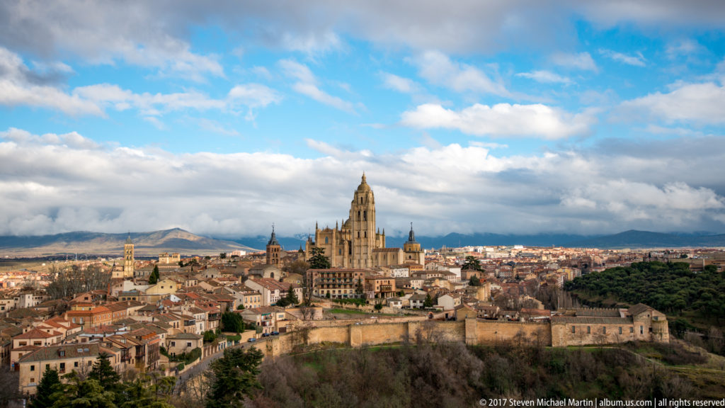 Elevated panorama of Segovia with cathedral tower above tiled roofs, old city walls, wooded slopes, distant hills, and layered clouds.