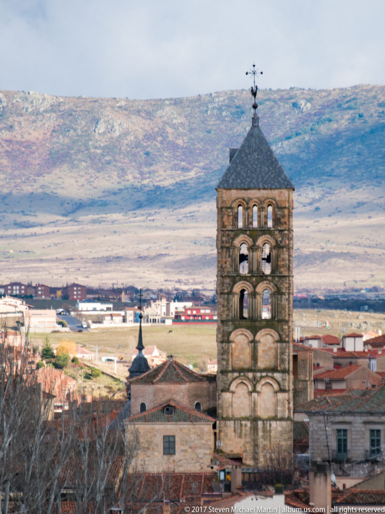 Square medieval bell tower with stacked round-arched openings, slate pyramidal roof, and weather vane against distant Segovia landscape.