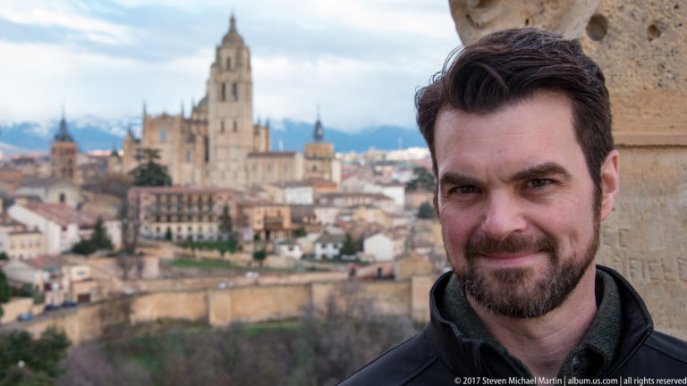 Bearded man in dark jacket at an overlook, with Segovia Cathedral and the historic city softly focused behind him.