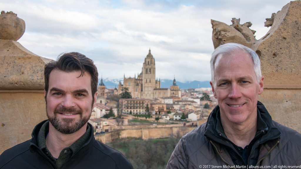 Two men posing at a stone overlook with Segovia Cathedral and the historic city visible behind them.