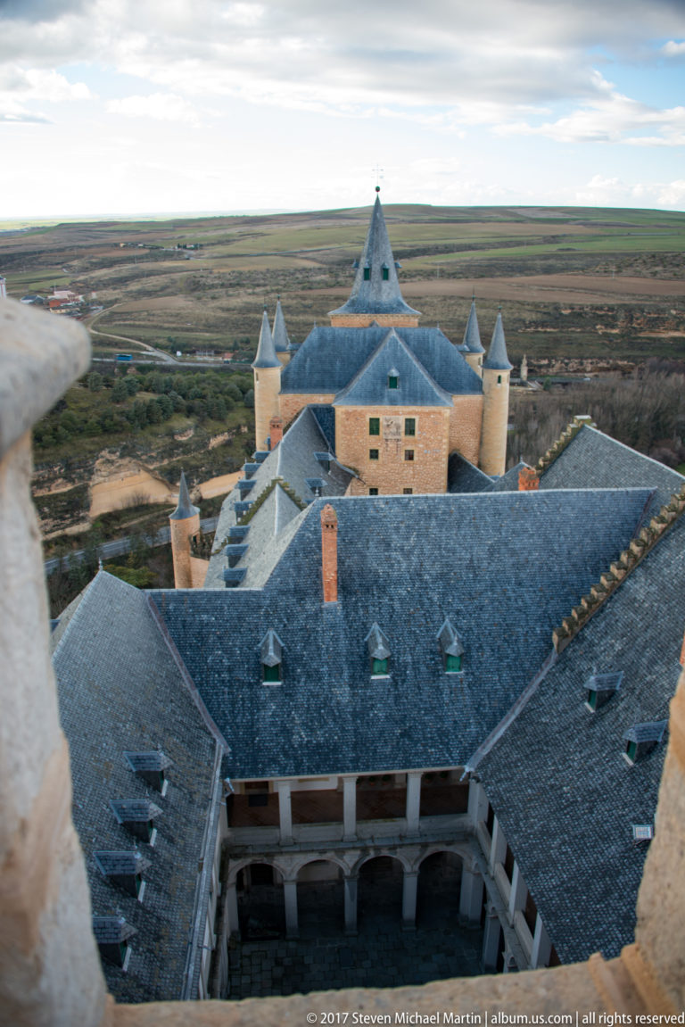 High view over slate roofs, dormers, chimneys, corner turrets, and an inner court of the Alcázar, with open countryside beyond.