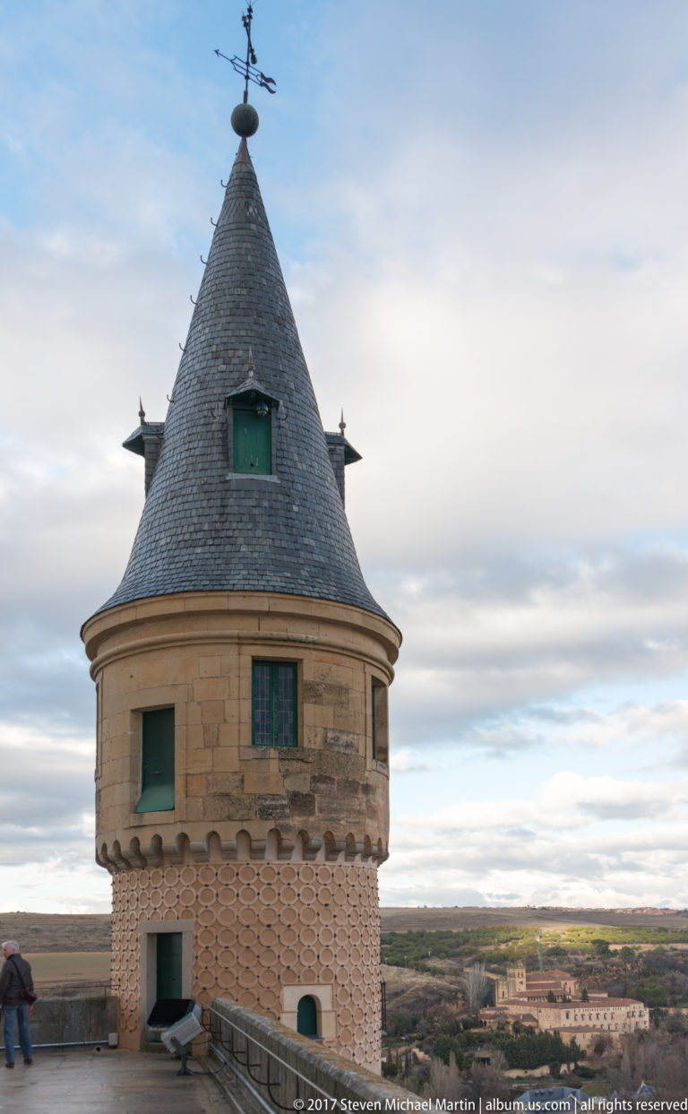 Round Alcázar tower with steep slate conical roof, weather vane, dormer opening, projecting stone band, and patterned masonry.