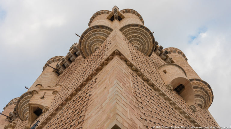 Extreme low-angle view up an Alcázar keep corner with projecting cylindrical turrets, corbels, and repeating circular wall patterns.