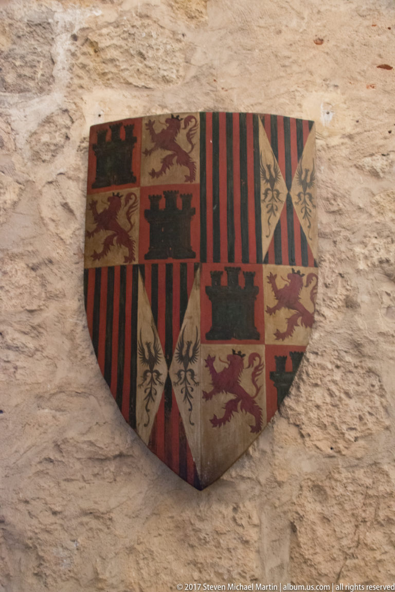 Painted quartered heraldic shield on a rough stone wall, with castles, lions, vertical stripes, and other colored emblems.