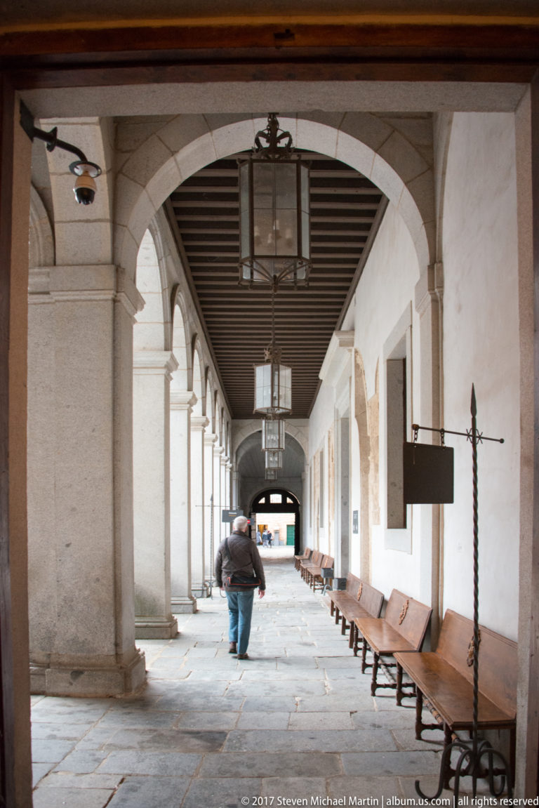Long Alcázar gallery with repeated stone arches, square piers, timber ceiling, hanging lanterns, benches, and one walking visitor.
