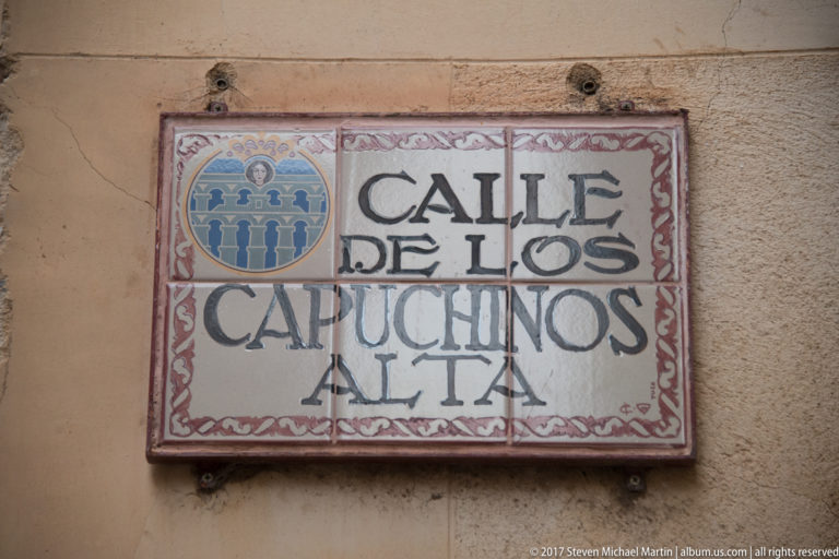 Ceramic street plaque reading Calle de los Capuchinos Alta, with black lettering, red border, and blue heraldic emblem.