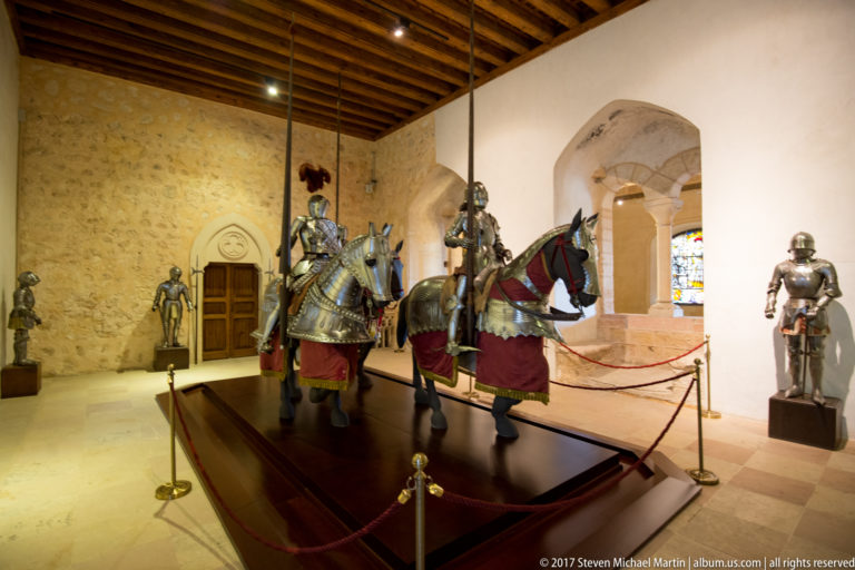 Museum gallery with armored rider on a caparisoned horse displayed on a platform among several freestanding suits of plate armor.