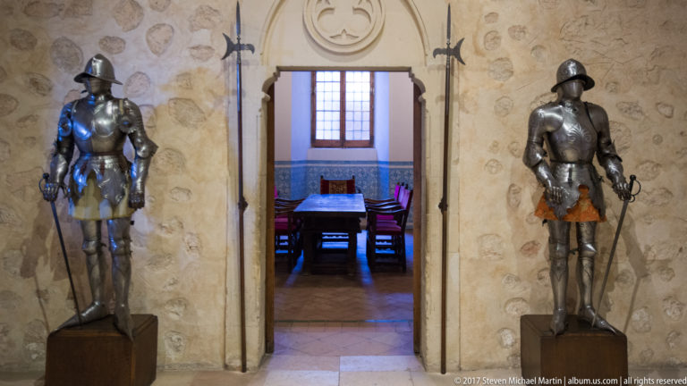 Two full suits of plate armor flank a stone doorway opening into a furnished room with a table and red chairs.