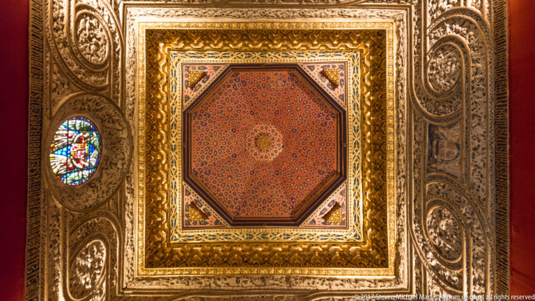 Upward view of a richly gilded geometric ceiling centered on an octagonal patterned panel with carved borders and small stained-glass openings.