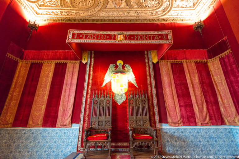 Red ceremonial room with two high-backed chairs beneath a large heraldic emblem, blue patterned tilework, and an ornate gilded ceiling.