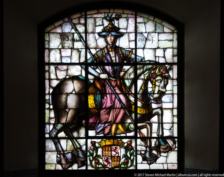Stained-glass window of an armored rider on horseback above a heraldic shield, set against a dark interior wall.