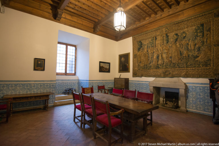 Historic room with a long wooden table, red chairs, large wall tapestry, stone fireplace, blue tilework, and timber ceiling.