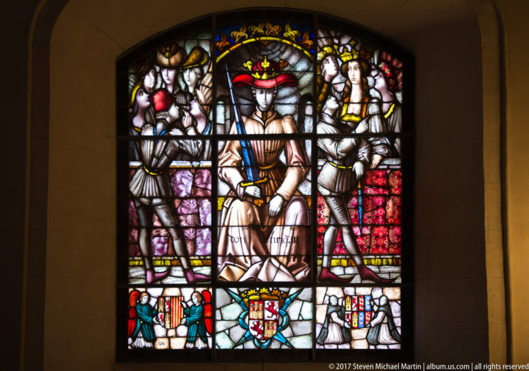 Stained-glass window with a seated female figure in a red hat, two armored attendants, and three heraldic shields below.