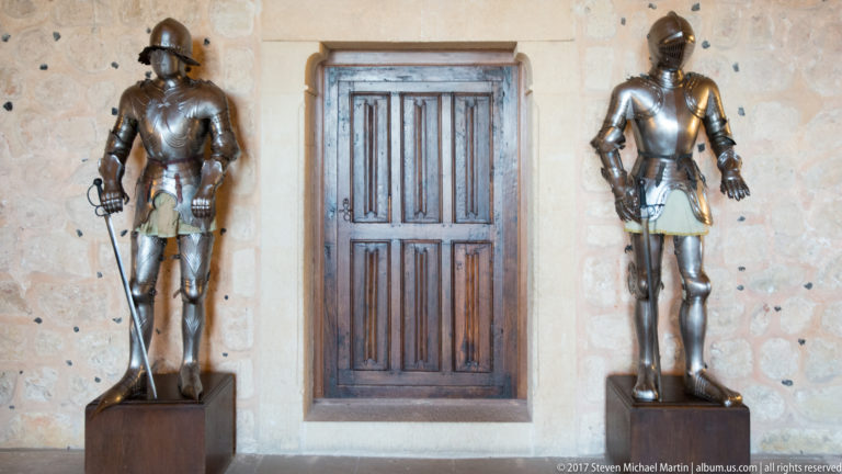 Two suits of polished plate armor stand on either side of a closed wooden door in a pale stone wall.