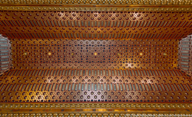 Decorative ceiling densely covered with repeated gold, red, blue, and black geometric patterns arranged in converging bands.